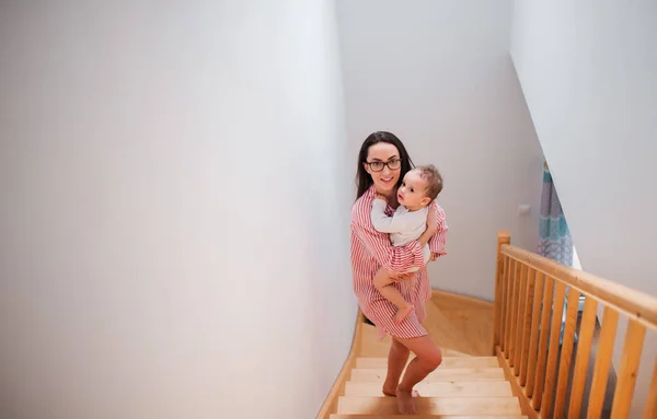 A young mother carrying small toddler son down the stairs indoors at home. — Stock Photo, Image