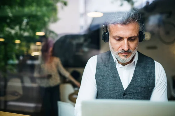 Mature man with headphones at the table in a cafe, using laptop. — Stock Photo, Image