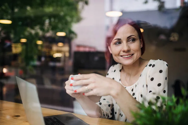 Um retrato de mulher com café sentado à mesa em um café, baleado através de vidro . — Fotografia de Stock