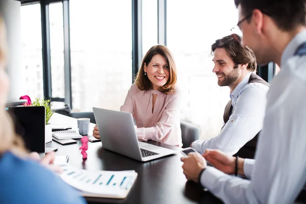 A group of young business people with laptop sitting in an office, talking. — Stock Photo, Image