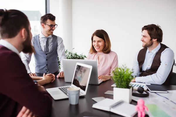 A group of young business people with laptop sitting in an office, talking. — Stock Photo, Image