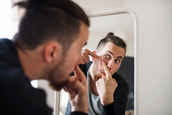 A young man indoors looking in the mirror, squeezing a pimple. — Stock Photo, Image