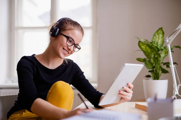 En ung kvinnlig elev som sitter vid bordet, använder hörlurar när man studerar. — Stockfoto