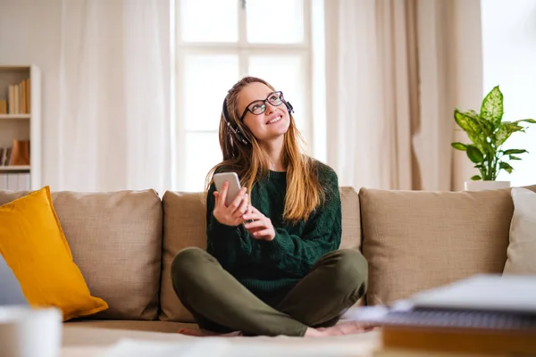 En ung kvinnlig elev som sitter vid bordet, använder hörlurar när man studerar. — Stockfoto