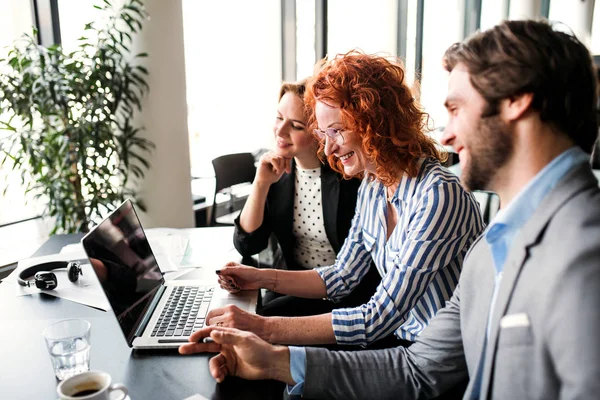 A group of young business people with laptop sitting in an office, talking. — Stock Photo, Image