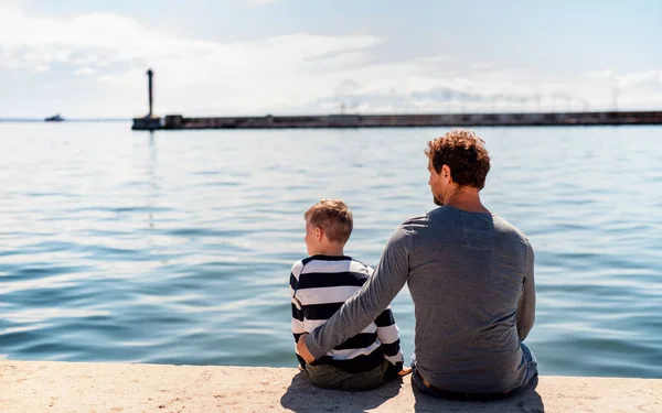 Vista posteriore del padre con il piccolo figlio seduto all'aperto in città in riva al mare, parlando . — Foto Stock