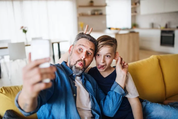 Pai com o filho pequeno sentado no sofá dentro de casa, sorrindo ao tomar selfie . — Fotografia de Stock