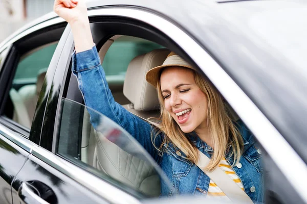 Young woman with hat sitting in car, listening to music and dancing. — Stock Photo, Image