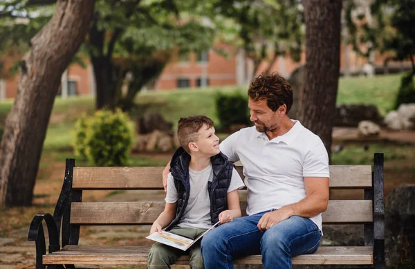 Padre con hijo sentado en el banco al aire libre en la ciudad, leyendo un libro . —  Fotos de Stock