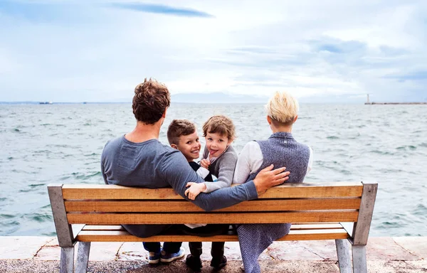 Νέα οικογένεια με δύο μικρά παιδιά κάθονται σε πάγκο σε εξωτερικούς χώρους στην παραλία. — Φωτογραφία Αρχείου