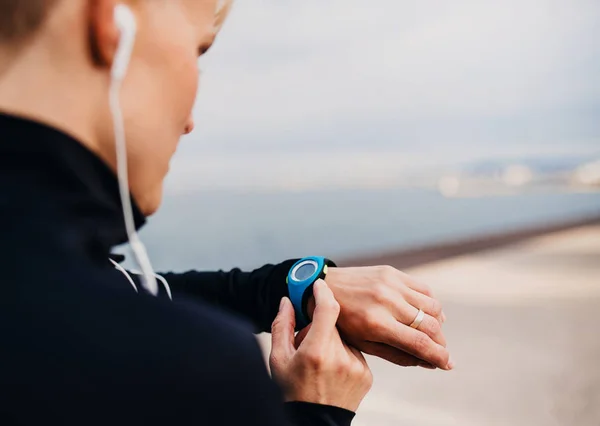 Eine junge Sportlerin mit Kopfhörern, die draußen am Strand steht und eine Smartwatch benutzt. — Stockfoto