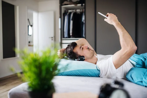 Jovem com fones de ouvido e smartphone na cama em casa, ouvindo música . — Fotografia de Stock