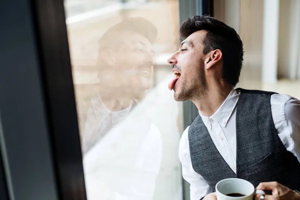 Kahvesi pencerenin yanında duran genç adam, dilini dışarı sokuyor.. — Stok fotoğraf