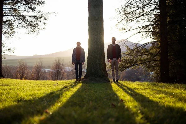 年长的父亲和他的儿子站在日落的树，看着相机. — 图库照片