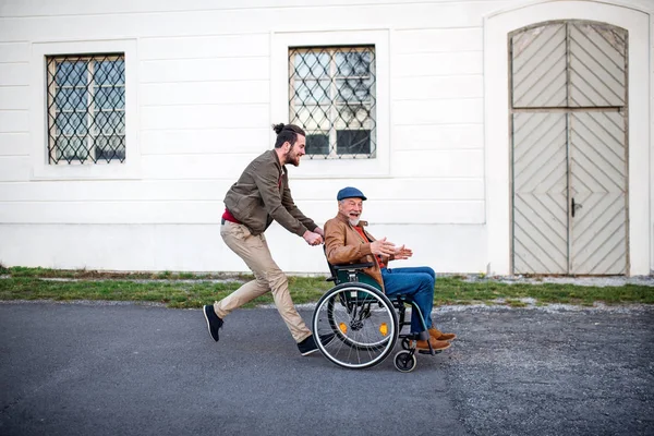 Jonge man en zijn senior vader in een rolstoel op een wandeling in de stad, plezier. — Stockfoto