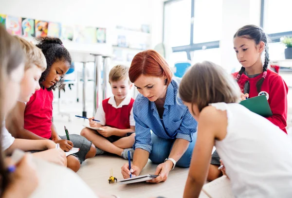 Μια ομάδα μικρών σχολικών παιδιών με δάσκαλο που κάθονται στο πάτωμα στην τάξη, μαθαίνοντας. — Φωτογραφία Αρχείου