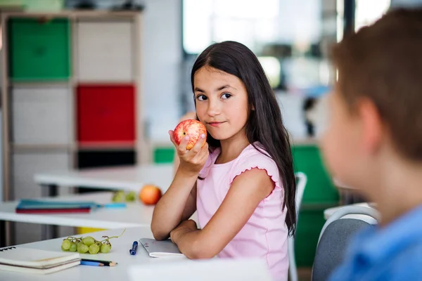 Uma menina da escola pequena sentada na mesa em sala de aula, comendo maçã . — Fotografia de Stock