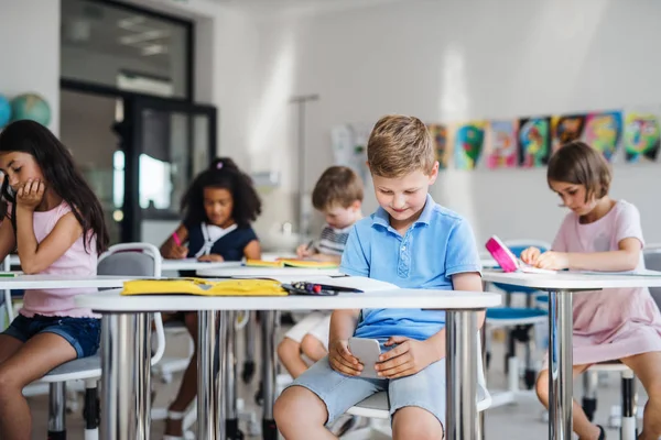 Un piccolo scolaretto con smartphone seduto alla scrivania in classe, che gioca . — Foto Stock