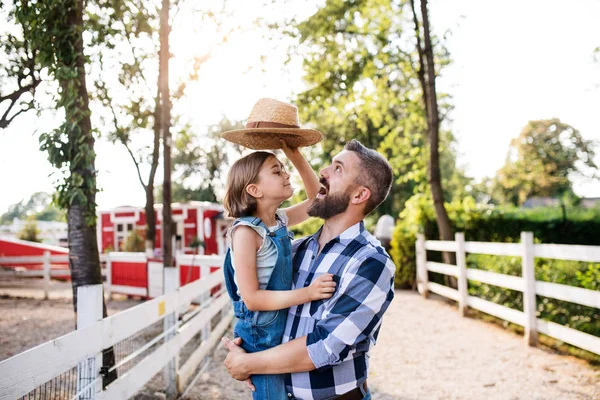 A father with small daughter standing outdoors on family farm, playing with hat. — Stock Photo, Image
