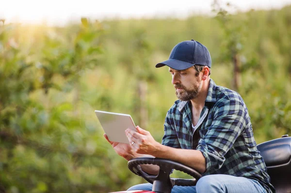 Um agricultor maduro com tablet sentado em mini trator ao ar livre no pomar . — Fotografia de Stock