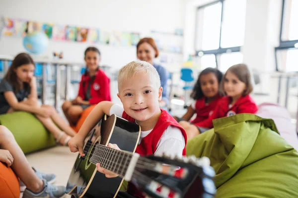 Un ragazzo down-sindrome con i bambini della scuola e insegnante seduto in classe, a suonare la chitarra . — Foto Stock