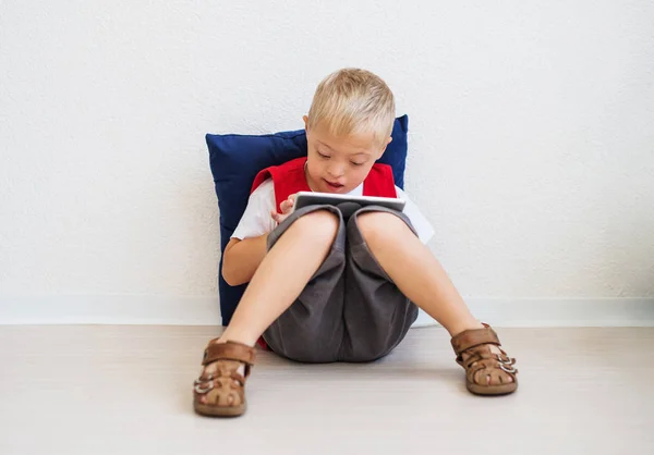 Un ritratto del ragazzo della scuola down-sindrome seduto sul pavimento, utilizzando tablet . — Foto Stock