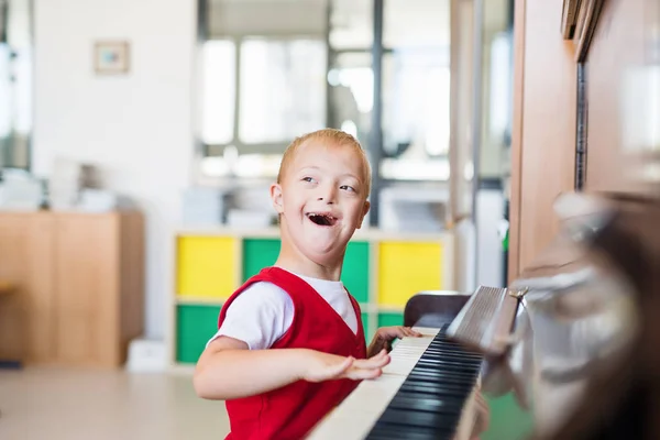 Um menino da escola com síndrome de down sentado na escola, tocando piano . — Fotografia de Stock