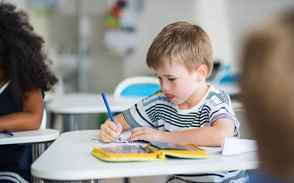 Ein kleiner Schuljunge sitzt am Schreibtisch im Klassenzimmer und schreibt. — Stockfoto