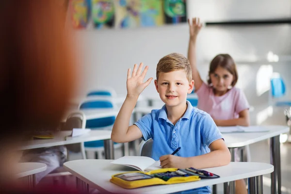 Crianças da escola sentadas na mesa em sala de aula sobre a lição, levantando as mãos . — Fotografia de Stock