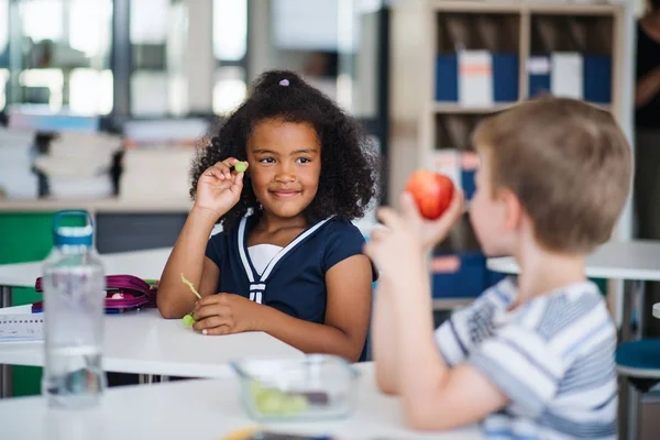 Crianças de escola pequena sentadas na mesa em sala de aula, comendo frutas . — Fotografia de Stock