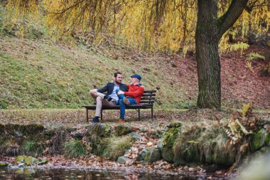 Kıdemli baba ve oğlu doğada göl kenarında bankta otururken, konuşuyor.
