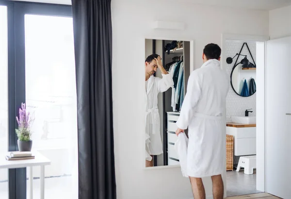 Jonge man op zoek in spiegel in de slaapkamer in de ochtend, dagelijkse routine. — Stockfoto