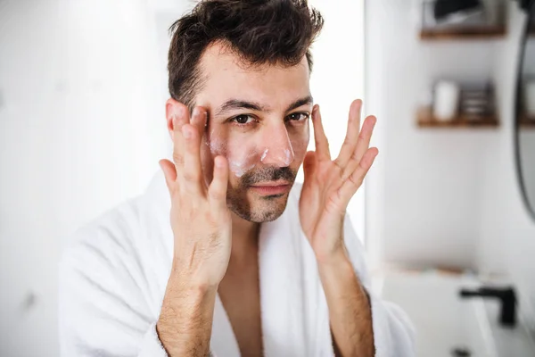 Young man putting cream on face in the bathroom in the morning, daily routine. — Stock Photo, Image