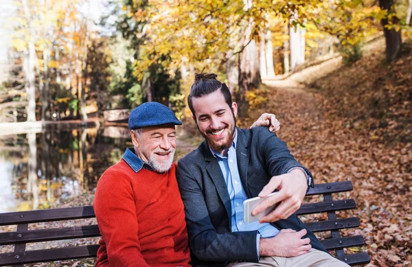 Pai sênior e seu filho sentado no banco na natureza, tomando selfie . — Fotografia de Stock