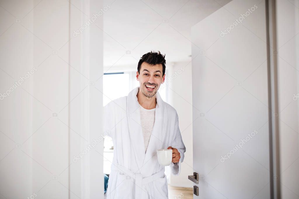 Front view of young man with coffee in bedroom in the morning.