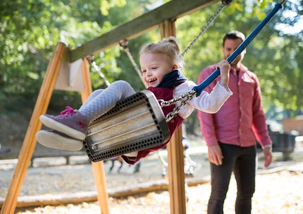 Una niña pequeña con un padre en un columpio en un parque infantil . —  Fotos de Stock