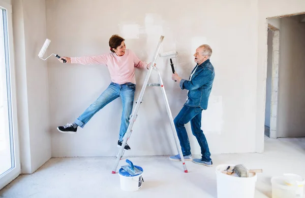 Senior paar schilderen muren in nieuw huis, plezier. Relocatie concept. — Stockfoto
