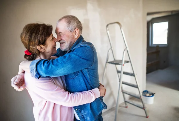 Un paio di anziani che dipingono muri in una nuova casa, abbracciati. Concetto di delocalizzazione . — Foto Stock