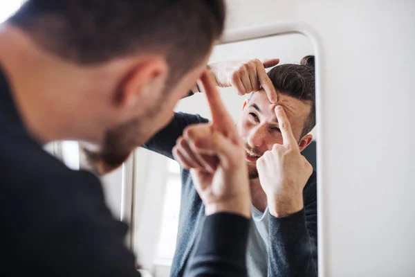 Un joven en el interior mirándose al espejo, apretando una espinilla . — Foto de Stock