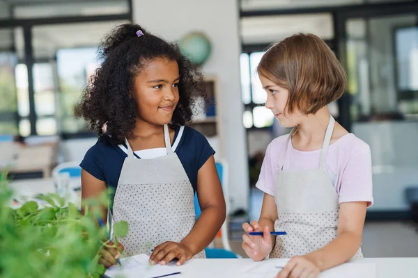 Piccoli scolari con grembiuli in piedi alla scrivania in classe, parlando . — Foto Stock