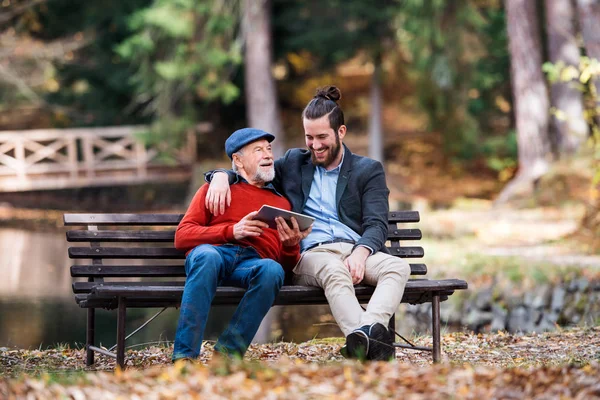 Senior far och hans son sitter på bänken vid sjön i naturen, med hjälp av Tablet. — Stockfoto