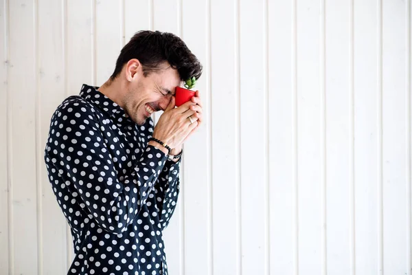 Frustrato giovane uomo in piedi contro parete di fondo in legno bianco, tenendo piccola pianta . — Foto Stock