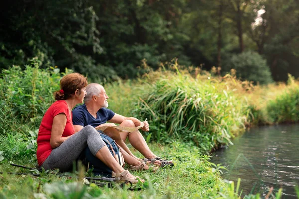 Senior toeristisch paar met kaart op een wandeling in de natuur, zittend aan het meer. — Stockfoto