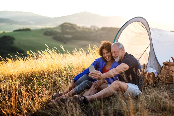 Senior turist par med smartphone sitter i naturen vid solnedgången, tar Selfie. — Stockfoto