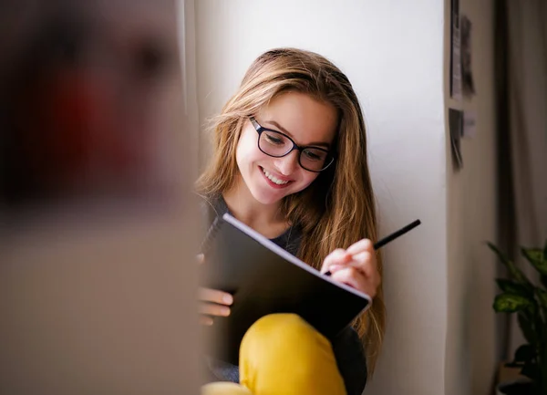 Een jonge vrouwelijke student met een oefenboek zittend op vensterbank, studeren. — Stockfoto