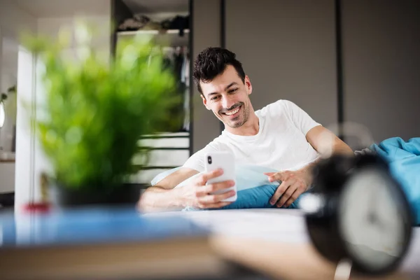 Ung man med smartphone i sängen hemma, textmeddelanden. — Stockfoto