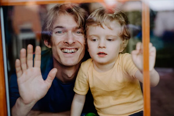 Un padre con un hijo pequeño en casa, mirando a la cámara. Disparo a través de vidrio . — Foto de Stock