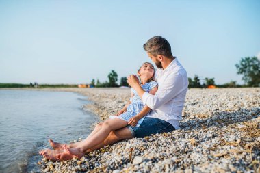 Göl kenarında oturan bir tatil baba ve küçük kızı yan görünümü.