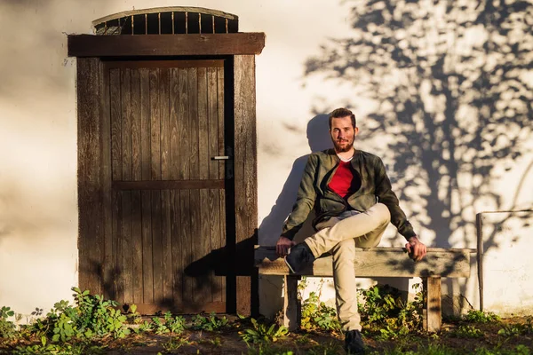 Lonely young man sitting on bench in front of old house, looking at camera.