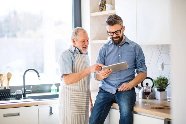 Adulte fils hipster et père aîné à l'intérieur dans la cuisine à la maison, en utilisant une tablette . — Photo